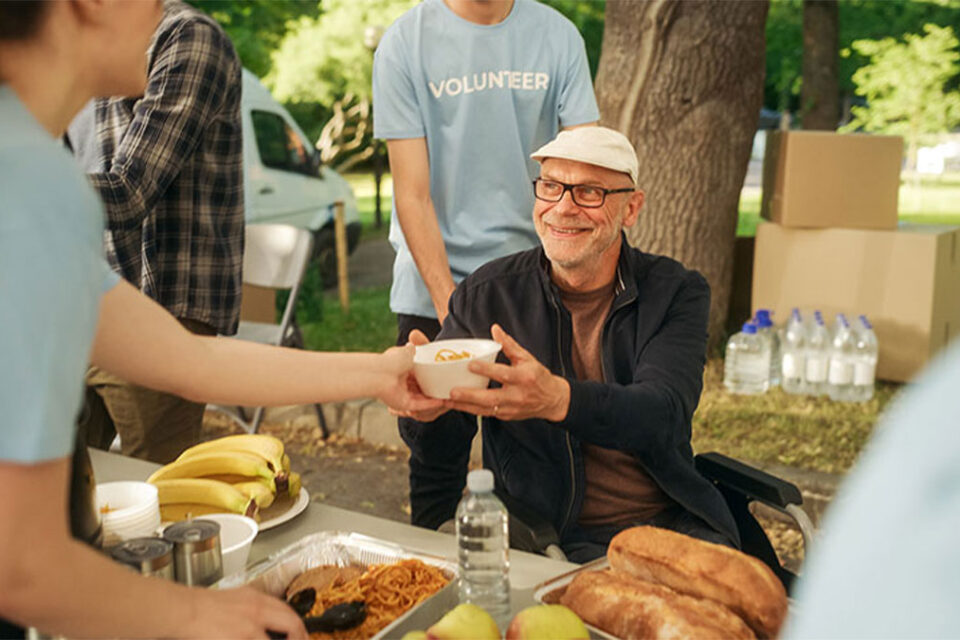 Man in a wheelchair accepting food from a volunteer during a food drive at a park